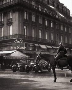 black and white photograph of people riding horses in front of an old building with cars parked on the street