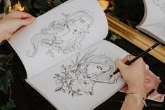 a person is drawing on an open book with flowers and leaves in the background, while another hand holds a pen