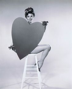 a black and white photo of a woman sitting on a stool holding a giant heart