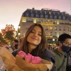 a woman holding a brown paper bag filled with pink donuts in front of a building