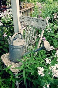 an old wooden chair sitting in the middle of some flowers and plants with a watering can on it