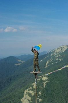 a man standing on top of a tall wooden pole holding a blue and yellow kite