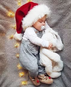 a baby wearing a santa hat and holding a stuffed animal in its mouth while laying on a bed