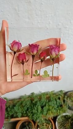 a hand holding a small glass block with pink flowers in it and green leaves on the inside