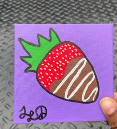 a hand holding up a purple piece of art with a chocolate covered strawberry on it