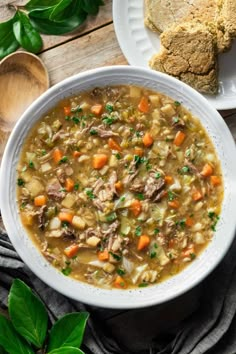 a white bowl filled with meat and vegetable soup on top of a wooden table next to bread