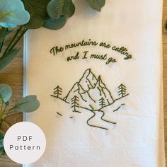 the mountains are calling and i must go embroidered tea towel on a wooden table with eucalyptus leaves