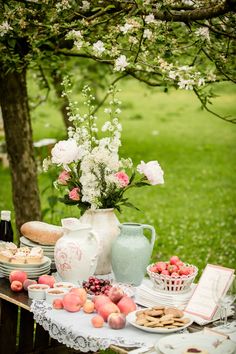 a table topped with plates and vases filled with fruit next to a tree covered in flowers