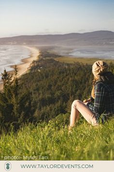 a woman sitting on top of a grass covered hill looking out at the ocean and mountains