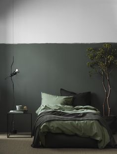 a bed with green sheets and pillows next to a small potted tree in the corner