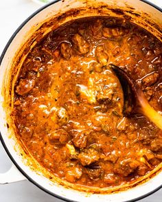 a pot filled with meat and sauce on top of a white table next to a wooden spoon