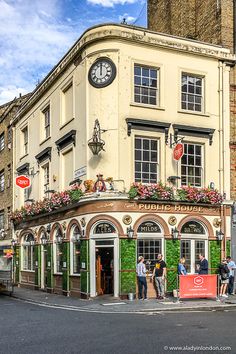 people are standing in front of a building with flowers on the outside and a clock mounted to it's side