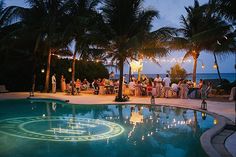 people are sitting at tables by the pool in front of palm trees with lights strung over them