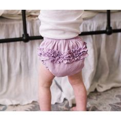 a baby standing on one foot in front of a crib and wearing purple ruffled blooming diapers