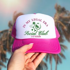 a person holding up a pink and white trucker hat that says in my legal era social club