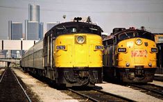 two yellow trains sitting side by side on train tracks in front of a city skyline