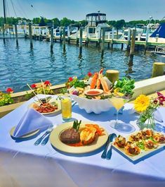 a table with food and drinks on it next to the water in front of boats