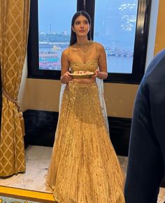 a woman standing in front of a window wearing a gold dress and holding a cake