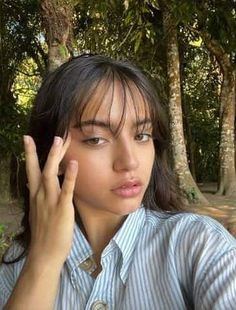 a young woman holding her hand up to her face in front of trees and dirt