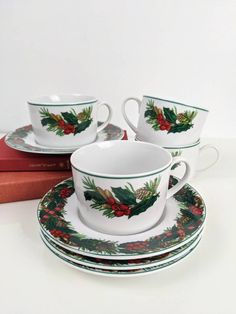 three christmas cups and saucers with holly decorations on them, sitting next to a book