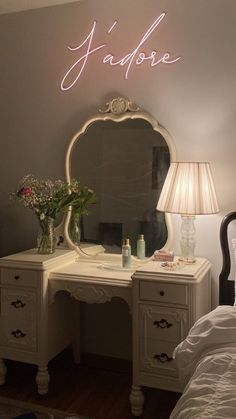 a white desk with a mirror, lamp and flowers on it next to a bed