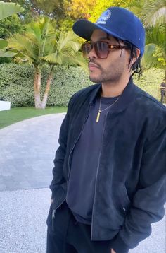 a man wearing a blue hat and sunglasses standing in front of a driveway with palm trees