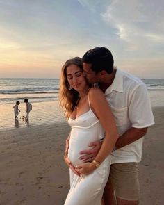 a pregnant woman hugging her husband on the beach at sunset with other people in the background