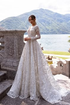 a woman in a wedding dress standing on some steps near the water with mountains behind her