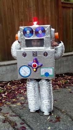 a robot made out of cds sitting on the ground next to a wooden fence with leaves around it