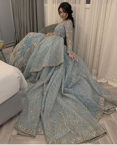 a woman in a blue dress sitting on a bed next to a gray couch and white curtains