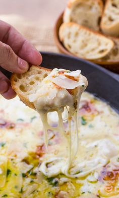 a hand holding a piece of bread over a bowl of soup with melted cheese on top