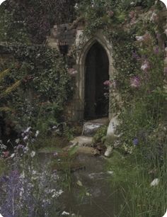 an old stone building surrounded by flowers and greenery with a small stream running through it