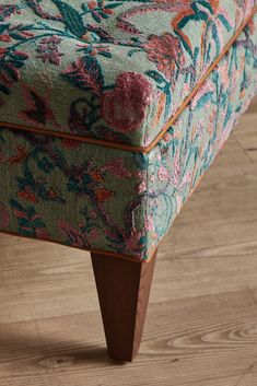 an upholstered footstool with floral fabric and wooden legs on a wood floor