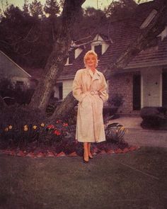 a woman standing in front of a house wearing a trench coat and matching heels with her hands on her hips