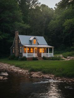 a small house sitting on top of a lush green field next to a river in front of a forest