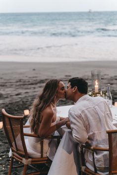 a man and woman sitting at a table with candles in front of them on the beach