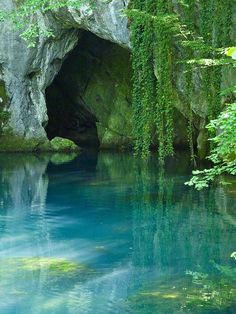 the water is blue and clear with green plants growing on it's sides, in front of a cave