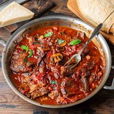 a pan filled with meat and tomato sauce next to bread on a wooden table,