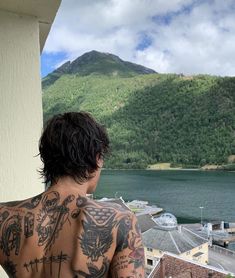 a man with tattoos on his back looking out at the water and mountains behind him