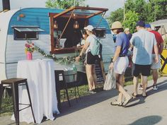 several people are standing outside near an old camper and some tables with white cloths on them