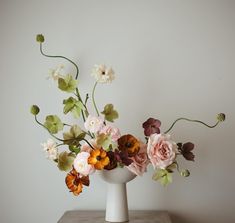 a white vase filled with lots of flowers on top of a wooden table next to a wall