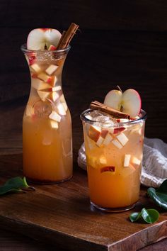 two glasses filled with apple cider on top of a wooden cutting board next to an apple