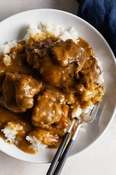 a white plate topped with meat covered in gravy and rice next to a fork