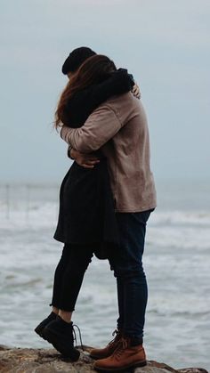 a man and woman standing on the rocks by the ocean hugging each other while looking at the water