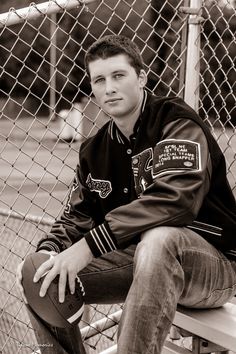 a young man sitting on a bench in front of a fence wearing a jacket and jeans