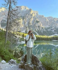 a person taking a photo with a camera near a mountain lake and pine trees in the background