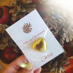 a person holding up a card with a heart shaped chocolate in the middle and pine cones behind it