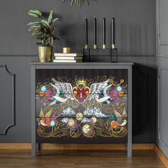 an artistically painted cabinet in the corner of a room with two candles and a potted plant