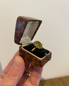 a person holding an open ring box with a green stone in it's middle