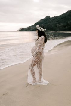 a pregnant woman standing on top of a sandy beach next to the ocean wearing a white dress